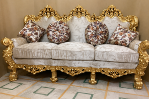 A premium handcrafted luxury sofa featuring an intricately carved antique gold solid wood frame with elegant floral upholstery. Complete with decorative round and square accent cushions, this masterpiece is designed to elevate luxury living rooms, hotels, villas, and palace-inspired interiors.